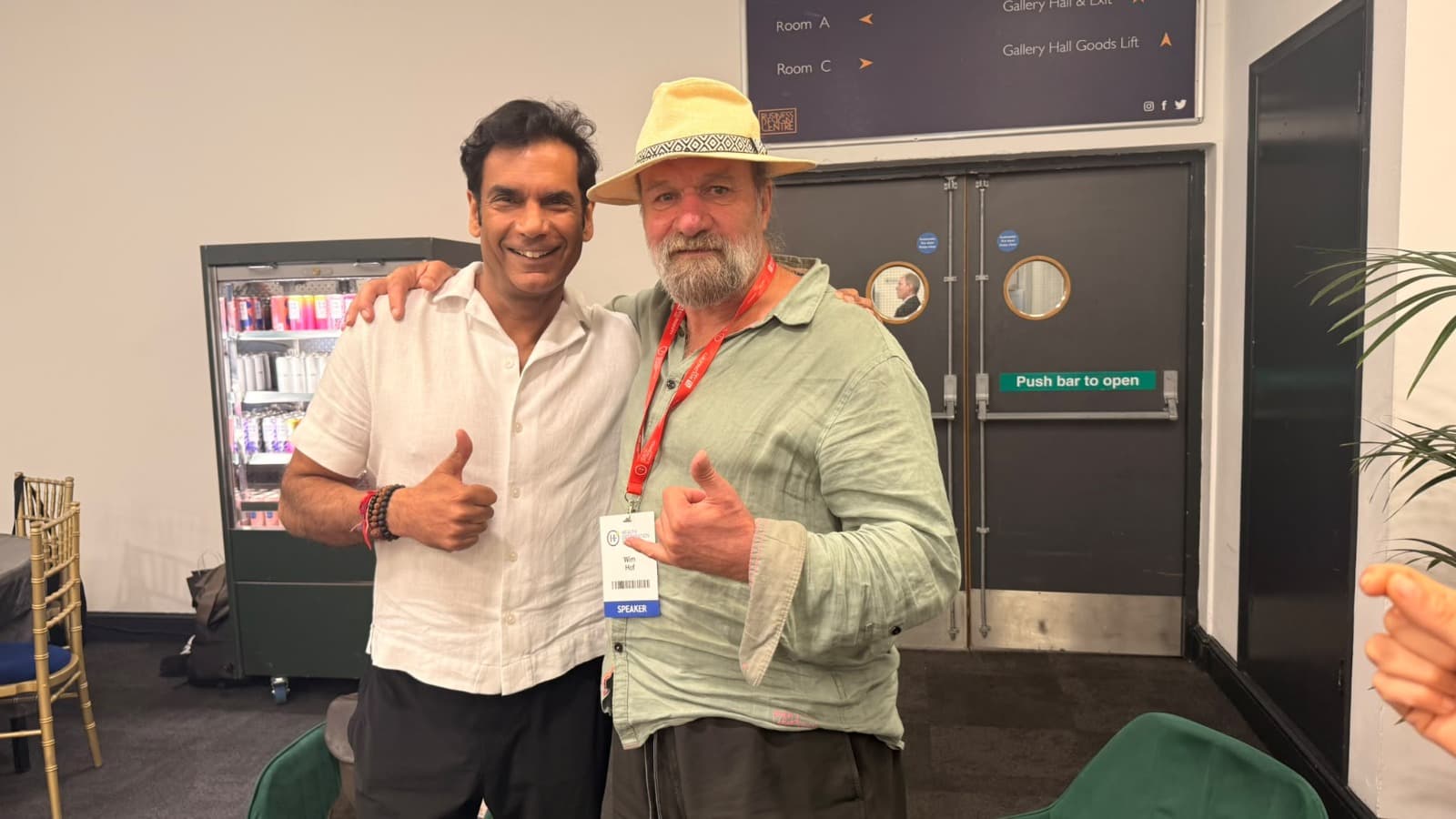 Two men smiling and giving thumbs-up at the Health Optimisation Summit, one in a white linen shirt and the other in a straw hat and green shirt with a red lanyard and speaker badge